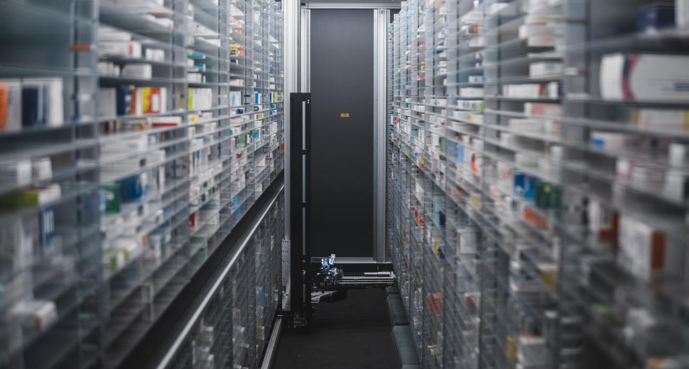 Narrow pharmacy aisle flanked by towering shelves stocked with medicine boxes. A pharmacy robot navigates through, ensuring efficient and precise medication retrieval.